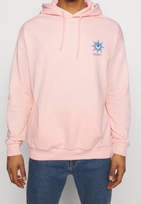 Sudadera rosa hecha de tela suave, con un bolsillo frontal, capucha con cordón y un gráfico de sol azul con la palabra "SERENIDAD".