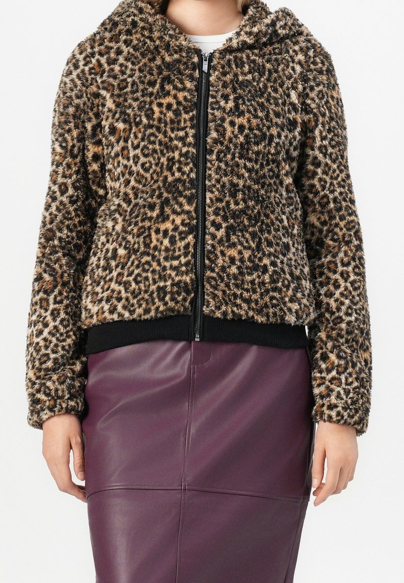 Personne portant une veste duveteuse zippée à imprimé léopard et une jupe en cuir violet foncé taille haute, mains détendues le long du corps.