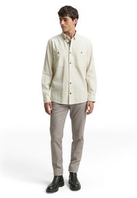 Cremefarbenes Hemd mit Knopfleiste, zwei Brusttaschen, schwarzen Knöpfen und strukturierter Stoffqualität, kombiniert mit hellgrauer Hose und schwarzen Stiefeletten.