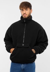 Pile nero con collo alto, mezza cerniera frontale e tasca a marsupio. Presenta una texture liscia e un logo discreto sulla tasca.