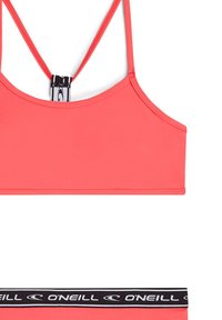 Reggiseno sportivo corallo realizzato in tessuto morbido; presenta spalline sottili e un elastico nero con logo decorativo alla base.