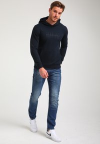 Sudadera navy con un bolsillo frontal y logotipo, combinada con unos jeans de mezclilla azules y zapatillas blancas con detalles en negro. Textura de tela suave.