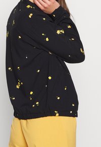 Černá květinová bunda s žlutými květy, elastický lem a manžety, funkční kapsy, doplněná světlými žlutými kalhotami.