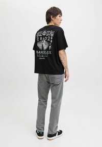 Ung mand med tatoveringer iført en sort grafisk T-shirt med teksten "RANDLUX ESTATES", grå jeans og sorte sneakers, står mod en hvid baggrund.