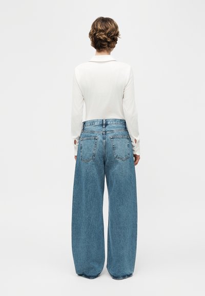 Άτομο που στέκεται με την πλάτη προς την κάμερα, φορώντας λευκή μπλούζα μακριάς μανίκας και ψηλόμεσα, φαρδιά τζιν παντελόνια ανοιχτού μπλε χρώματος, μπροστά σε απλό φόντο.