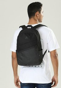 Zaino nero realizzato in materiale resistente, dotato di spallacci imbottiti, una tasca frontale con zip e una tasca laterale in rete per bottiglie d'acqua.