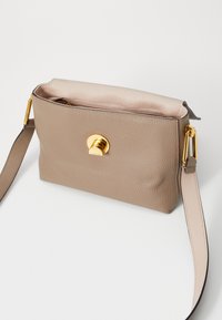 Sac en cuir beige porté travers avec une finition texturée, doté d'un fermoir circulaire en or et d'une bandoulière plate pour un transport facile.