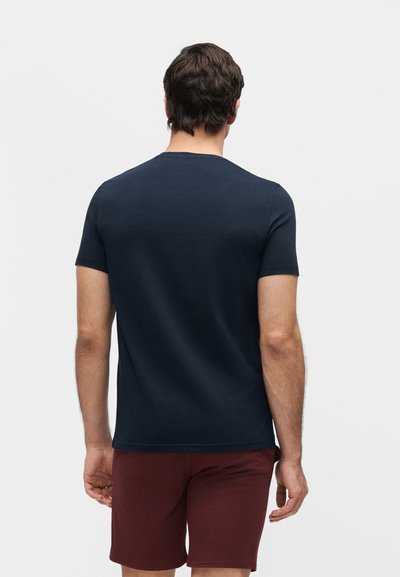 Άνδρας που στέκεται με την πλάτη στην κάμερα, φοράει μπλε ναυτικού κοντομάνικο μπλουζάκι και μπορντό σορτς μπροστά σε καθημερινό φωτεινό φόντο.