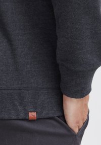 Dunkelgrauer Sweatshirt mit gerippten Bündchen und Saum, versehen mit einem kleinen braunen Markenetikett an der unteren linken Seite. Weicher Texture und lässige Passform.