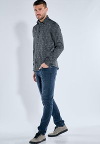 Mann steht da, trägt ein dunkel gemustertes Langarmhemd, blaue Jeans und beigefarbene Sneaker, mit einer Hand in der Tasche.