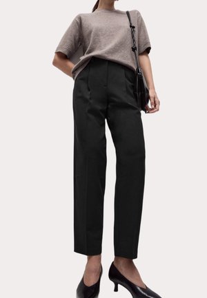 Vrouw draagt een losvallende taupe kortemouwentrui, hooggetailleerde zwarte wijde broek, zwarte hakken en draagt een zwarte schoudertas.