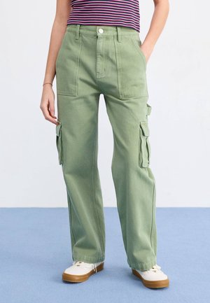 Donna che indossa pantaloni cargo verde chiaro a gamba larga con grandi tasche anteriori e laterali, sneakers bianche e una camicia a righe.