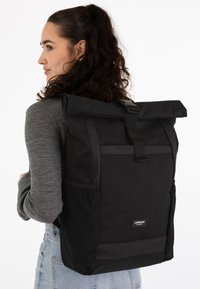 Zaino nero con chiusura arrotolabile, realizzato in tessuto resistente, dotato di spallacci regolabili, tasca frontale e tasche laterali in rete per ulteriore spazio di archiviazione.