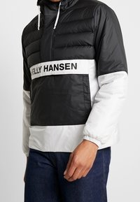 Giacca puffer bianca e nera con design a mezza zip, caratterizzata da una fascia bianca con il testo "HELLY HANSEN" sul petto e polsini elasticizzati.