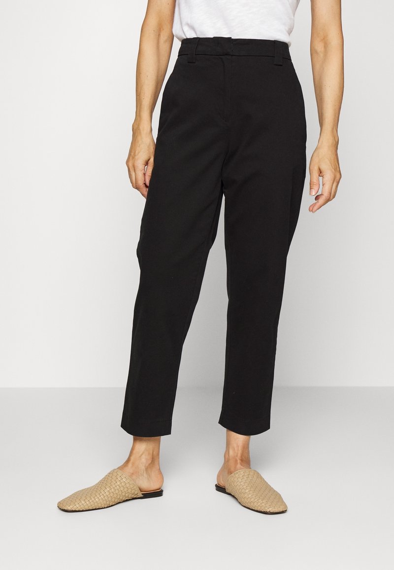 Femme portant un pantalon court taille haute noir, un haut blanc et des chaussures beige tissées à enfiler, debout devant un fond blanc uni.
