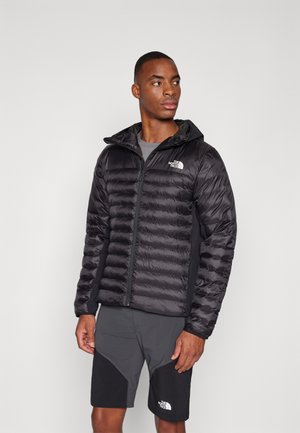 Zwarte gewatteerde jas met een capuchon, voorzien van een gestikte patroon, ritssluiting aan de voorzijde en logo. Gecombineerd met grijze shorts en een grijze binnenvoering.