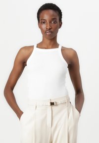 Weißes, geripptes Tanktop mit hohem Kragen, kombiniert mit beigen, hoch taillierten Hosen mit einem Gürtel und seitlichen Taschen.