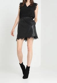Schwarzes ärmelloses Top mit Spitzenbesatz, kombiniert mit einem schwarzen Lederrock mit Spitzensaum. Schwarze Knöchelstiefel runden den Look ab.
