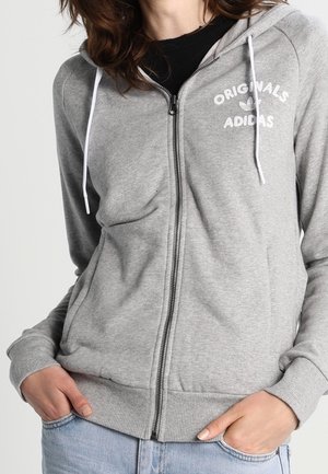 Person iført en grå Adidas Originals hættetrøje med lynlås og hvide snore, med hænderne i lyseblå jeanslommer.
