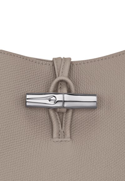 Beige strukturierte Lederhandtasche oben mit einem silbernen metallischen Verschluss und dekorativen Schnüren. Der Verschluss weist ein ovales Ausschnittdetail auf.