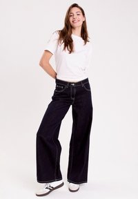 Mørkeblå bredbenede jeans har kontrastfarvet hvid søm, en knaplukning foran og fem lommer, som er stylet med en simpel hvid t-shirt.