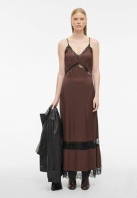 Bruine satijnen jurk met zwarte kantaccenten, V-hals en uitsnijdingen. Model houdt een zwarte leren jas vast en draagt bruine enkellaarzen.