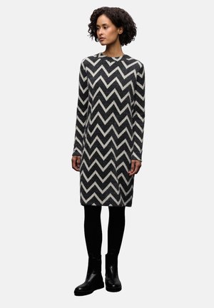 Musta-valkoinen chevron-kuvioinen neulemekko, jossa pitkät hihat, pyöreä pääntie, suora istuvuus ja pehmeä koostumus, yhdistettynä mustiin nilkkureihin.