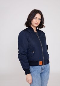 Marineblå bomberjakke med lynlåsfortil, ribbede manchetter og et orange mærke i taljen. Glat, let materiale med sidelommer.