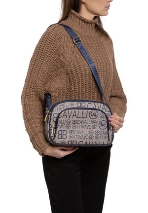 Beige schoudertas met marineblauwe accenten, met een logo-patroon, gouden hardware en een verstelbare schouderband. Zachte textuur, gestructureerde vorm.