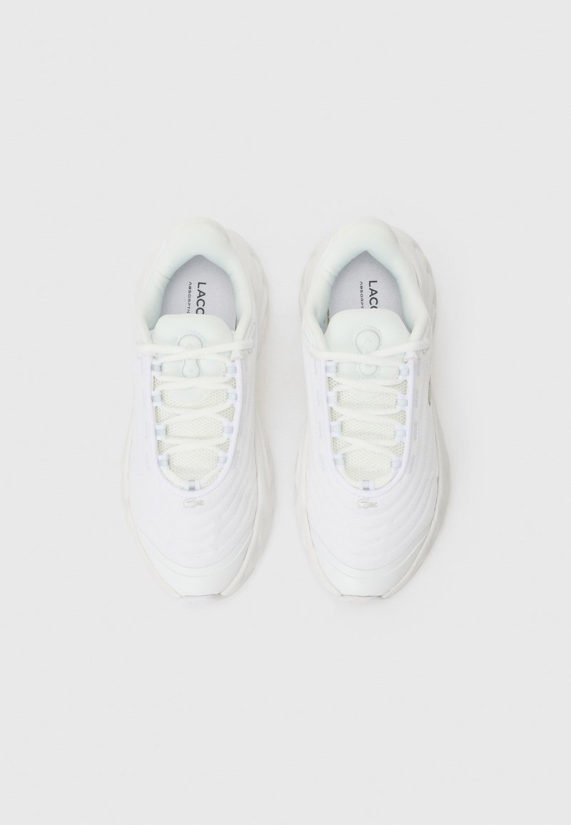 Zapatillas blancas con parte superior texturizada, que presentan cierre de cordones, puntera redonda y logo visible. Material suave y diseño de suela curvada.