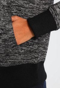 Zwart en grijs gestreepte sweater met geribbelde manchetten, voorzien van een zijzak en een textuurstof. Close-up van een hand in de zak.