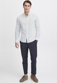 Camisa de botones a rayas azul claro y blanca, pantalones oscuros de corte slim y zapatos de ante beige. Diseño simple con líneas limpias.