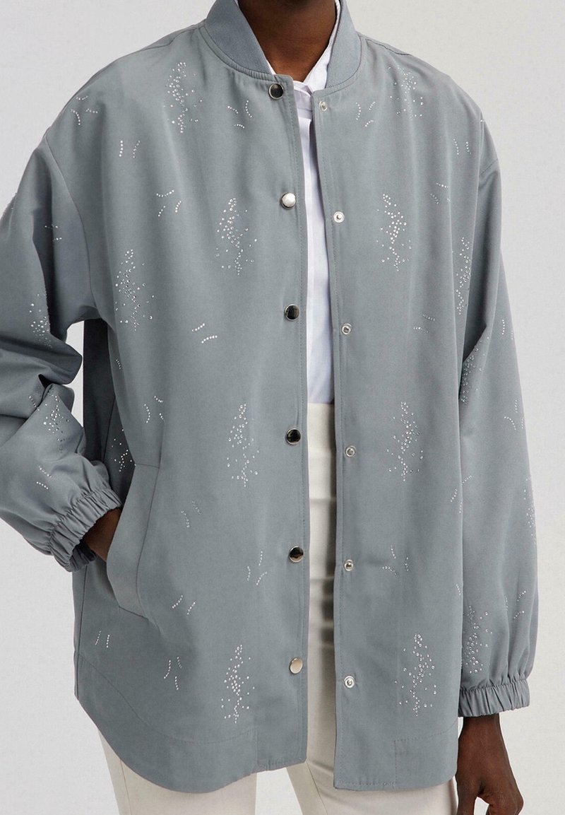 Grå jakke med en blød tekstur, der har knaplukning i front, elastiske manchetter og dekorative sølvforziehninger i forskellige mønstre.