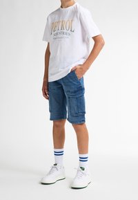 Bílá tričko s logem "PETROL", modré cargo šortky s několika kapsami, bílé tenisky s zelenými akcenty a pruhované bílé ponožky.