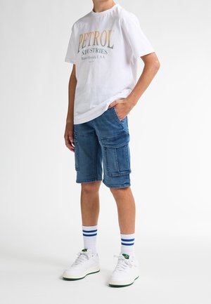 Bílá tričko s logem "PETROL", modré cargo šortky s několika kapsami, bílé tenisky s zelenými akcenty a pruhované bílé ponožky.