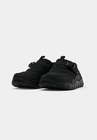 Pár černých slip-on polstrovaných bot s tlustými podrážkami a nastavitelnými bočními popruhy na jednobarevném bílém pozadí.