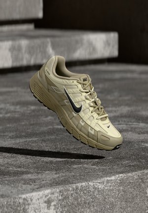 Beige Nike running shoe with black swoosh mid-air above concrete steps, showing textured sole and mesh fabric upper.