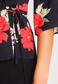 Blouse fleurie avec un fond bleu marine foncé orné de grandes fleurs rouges et roses. Elle a des manches courtes et une taille cintrée avec un lien pour un look ajusté.
