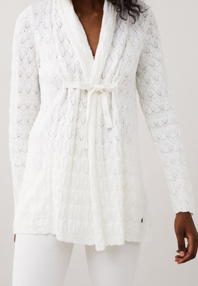 Cardigan bianco lavorato a maglia con motivo a diamante, maniche lunghe e chiusura a nodo frontale. Texture morbida con una vestibilità leggermente oversize.