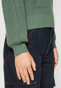 Primo piano di una persona che indossa un maglione di lana verde e jeans in denim blu scuro, con la mano sinistra appoggiata accanto a sé.