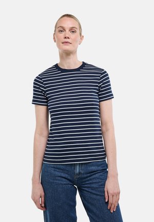 Femme aux cheveux blonds portant un t-shirt à manches courtes rayé bleu marine et blanc et un jean bleu, debout devant un fond uni.