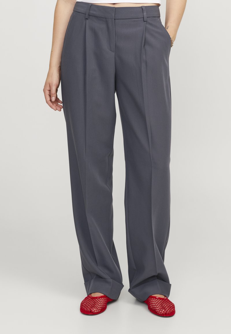 Pantalones grises, ajustados, de pierna ancha con pliegues verticales y bolsillos inclinados, combinados con zapatos de malla rojos. Acabado de tela suave.