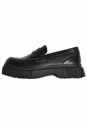 Mocassino in pelle nera con suola spessa in gomma tipo carrarmato e una larga fascia sulla tomaia, mostrato di profilo su sfondo bianco.
