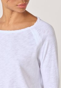 Kvinna klädd i en vit tröja med långa ärmar och en bred halslinning med råa kanter, som visar nacke, axel och en del av ansiktet vänt åt sidan.