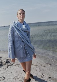 Ljusblå kabelstickad cardigan med knappar, matchad med en scarf. Bärs över en figurnära kjol. Bruna ankelboots på sandig strand.