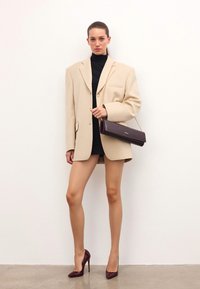 Kobieta w oversizowej beżowej marynarce na czarnej mini sukience, trzymająca ciemnobrązową torebkę kopertową i burgundowe szpilki, stojąca przed białą ścianą.