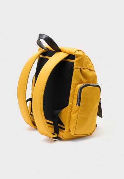 Sac à dos jaune avec bretelles rembourrées, panneau arrière en maille noire et une petite poche frontale zippée sur un fond clair.
