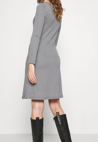 Mustat ja valkoiset pitkähihainen mekko, jossa on geometrinen kuvio, istuva yläosa, levenevä hame ja sileä pinta, yhdistettynä polven yli ulottuviin mustiin saappaisiin.