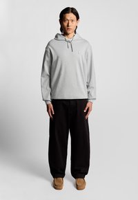 Felpa grigia chiara con tasca frontale, cappuccio con cordino e piccolo logo. Indossata con pantaloni neri a gamba larga e scarpe slip-on beige.