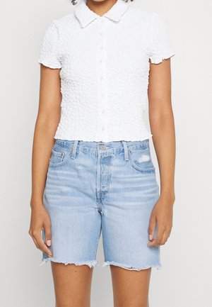 Femme portant une chemise blanche texturée à manches courtes boutonnée et un short en jean bleu clair taille haute avec des ourlets effilochés.